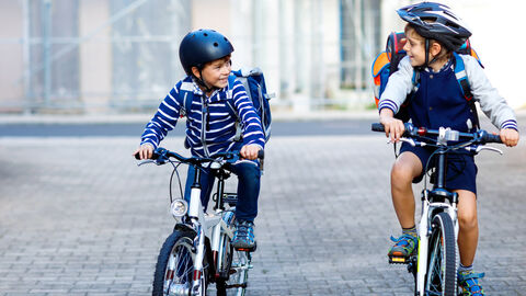 Auf dem Schulweg Zwei Kinder fahren lachend nebeneinander auf Fahrrädern. Sie haben Helme und Schulranzen auf.