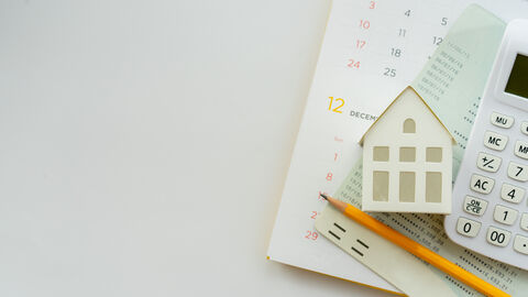 Ein Kalender, Unterlagen, ein Taschenrechner, ein kleines Häuschen aus Papier und ein Bleistift liegen auf einem Tisch.