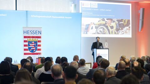 Verkehrsminister Mansoori steht auf einer Bühne vor Publikum am Rednerpult. Hinter ihm ein Banner mit dem Hessen-Wappen und dem Schriftzug "Arbeitgemeinschaft Nahmobilität Hessen"