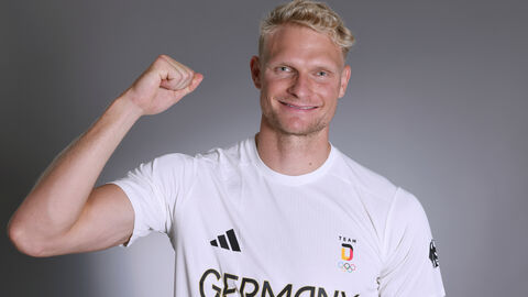 Ein blonder Mann mit weißem T-Shirt streckt den arm hoch und hat einen begeisterten Gesichstausdruck