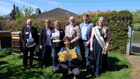 Sechs Personen stehen auf einer Wiese vor mehreren Bienenstöcken. Im Vordergrund kniet ein Mann und hält eine Wabe in die Kamera. 