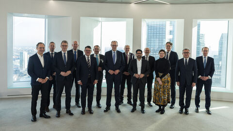 Eine Gruppe von Menschen stehen in einem Raum in einem Hochhaus. Im Hintergrund ist ein anderes Hochhaus aus dem Fenster zu sehen.
