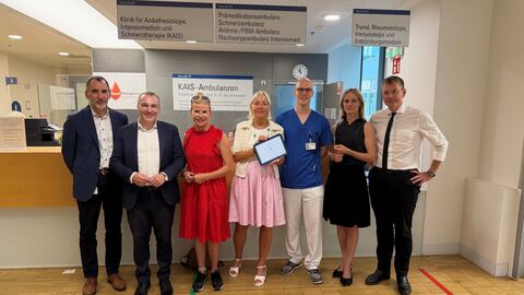 Eine Gruppe von sieben Personen steht lächelnd nebeneinander im Empfangsbereich einer Klinik (KAIS-Ambulanzen). Eine Frau in der Mitte hält ein Tablet hoch. Im Hintergrund sind Hinweisschilder für verschiedene medizinische Abteilungen zu sehen.