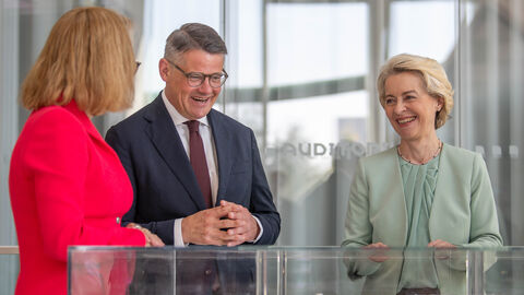 Unternehmensbesuch Merck Ministerpräsident Rhein und Ursula von der Leyen stehen gemeinsam vor einem Kasten mit einem Modell. Sie unterhalten sich mit einer dritten Person.