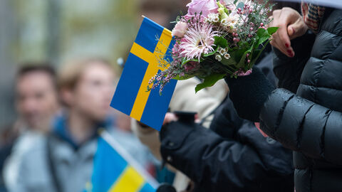 Nahaufnahme einer schwedischen Flagge in der Hand einer Person. Dazu hält diese Person auch noch einen Blumenstrauß in der Hand.
