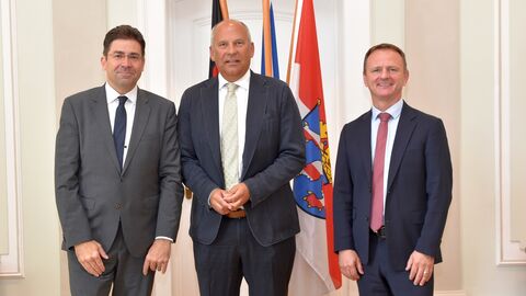 Regierungspräsident Prof. Dr. Jan Hilligardt , Innenminister Roman Poseck und Vizepräsident Dr. Stefan Fuhrmann (v.l.n.r.) stehen nebeneinander, im Hintergrund stehen Flaggen.