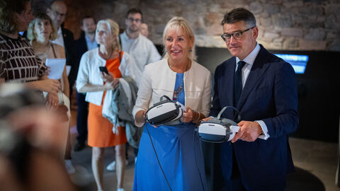 Digitalministerin Sinemus und Ministerpräsident Rhein halten VR-Brillen in Kamera. Im Hintergrund sieht man weitere Menschen.