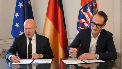 Vertragsunterzeichnung GEMA Zwei Personen sitzen an einem Tisch und unterzeichnen Unterlagen. Im Hintergrund sind die Flaggen von Europa, Deutschland und Hessen aufgestellt.