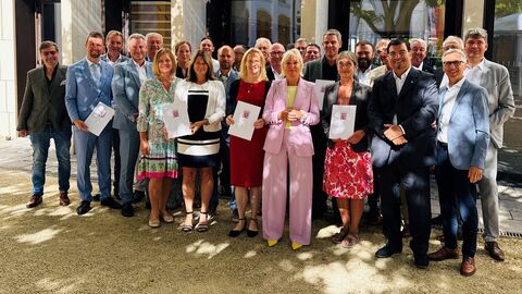Digitalministerin Prof. Dr. Kristina Sinemus hat an 13 Kommunen Förderbescheide zur Installation eines Starkregenfrühalarmsystems überreicht.  Dr. Sinemus steht in einer Reihe mit mehreren festlich gekleideten Menschen und hält einen Förderbescheid in den Händen.
