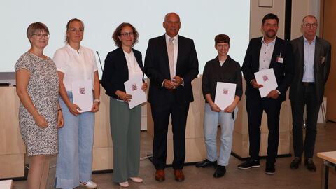Förderperiode des Landesprogramms „Hessen – aktiv für Demokratie und gegen Extremismus“  Mehrere Personen stehen vor einer Leinwand. Einige halten eine Mappe mit dem Hessenwappen darauf.