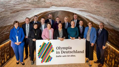 Eine Gruppe von 15 Personen steht in einem Kellergewölbe - vor Ihnen ein Schild mit der Aufschrift "Olympia in Deutschland".