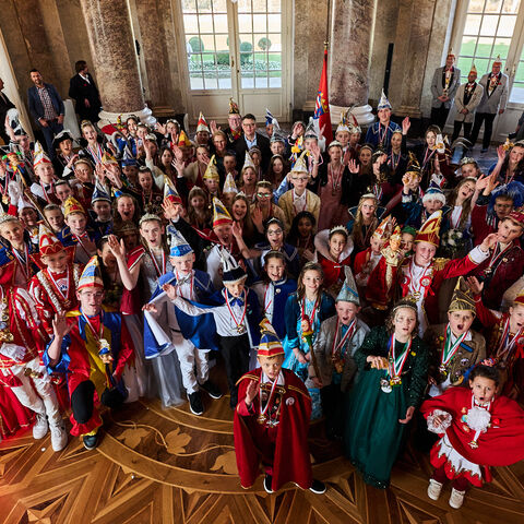 Närrisches Wimmelbild mit Ministerpräsidenten 39 Kinderprinzenpaare stehen jubelnd in einem Saal, in ihrer Mitte der hessische Ministerpräsident.